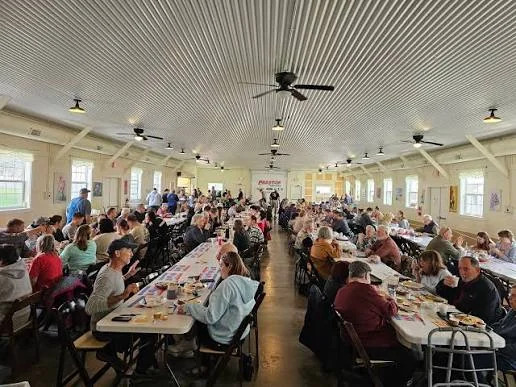 A community gathering fills the village event hall, with guests seated at long tables.